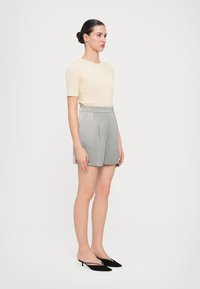 Haut beige à manches courtes associé à un short gris taille haute. Le modèle porte des escarpins à bouts pointus noirs. Design simple, tissu lisse.