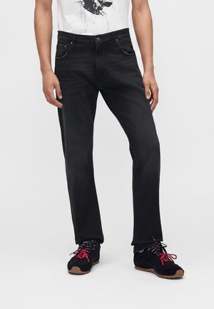 Homme portant un jean noir et des baskets noires avec des lacets rouges et noirs sur un fond blanc uni.