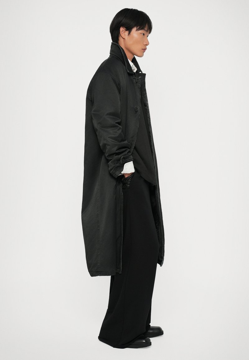 Manto lungo nero con colletto, chiusura frontale con bottoni e tasche laterali, indossato sopra un abbigliamento nero ampio e scarpe nere. Texture liscia.