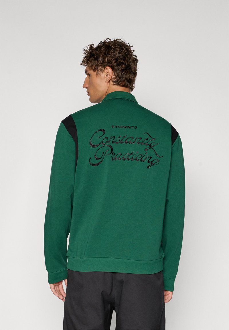Grønn hettegenser med svarte skulderdetaljer, med brodert tekst "STUDENTER Øver Konstant" på baksiden. Blandingsmateriale av bomull.