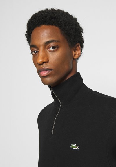 Le pull à col zippé noir présente une texture côtelée, une coupe slim et un logo de crocodile brodé en vert sur la poitrine gauche.