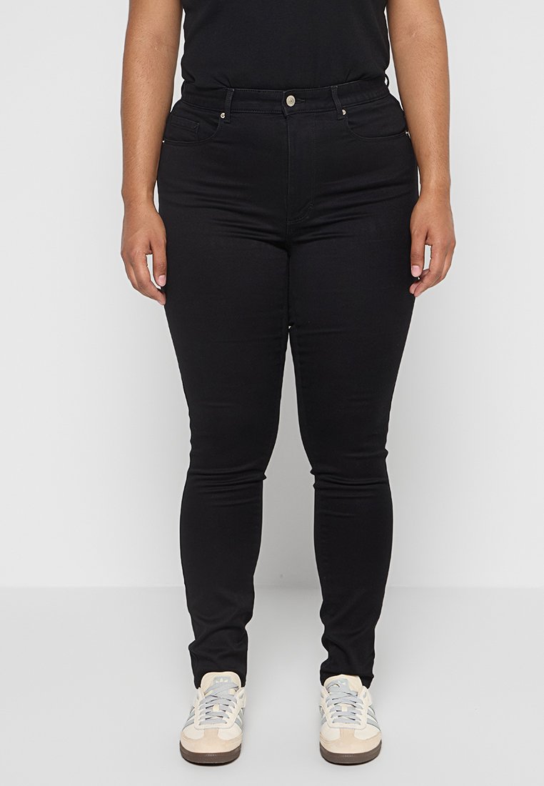 Schwarze dehnbare Jeans mit schmaler Passform, fünf Taschen, vorderer Knopfverschluss und dezenten Nähdetails, kombiniert mit hellen Sneakers.