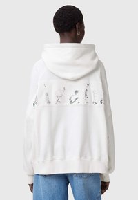 Sudadera crema con un gran logotipo bordado en la parte posterior. Presenta una capucha con cordones y puños acanalados. Combinada con jeans azules.