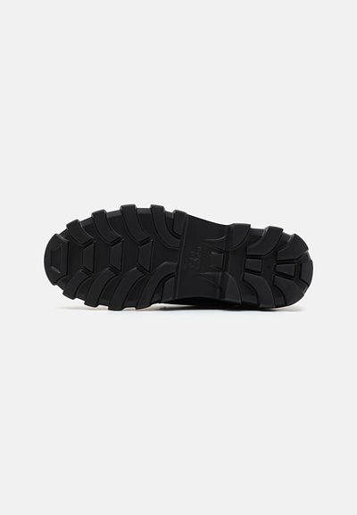 Semelle de chaussure en caoutchouc noir avec un motif rainuré, comportant des cannelures pour une adhérence et une durabilité accrues. Marquage discret près du talon.
