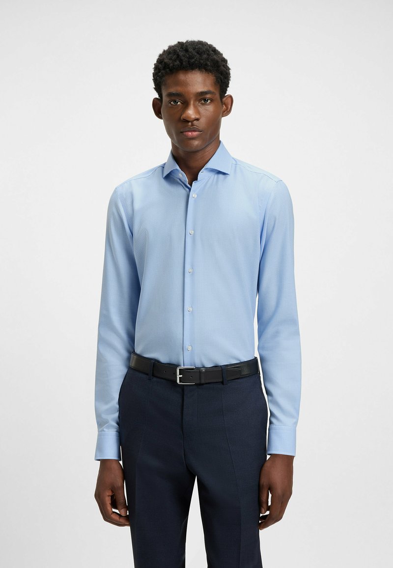 Vaaleansininen, pitkähihaisella paidalla, jossa on hienovarainen ruudullinen kuvio, nappikaulus ja istuva malli, yhdistettynä tummansinisiin housuihin ja vyöhön.