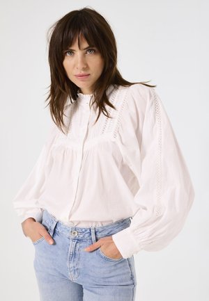 Witte blouse met tekstuelle schouderpartij en opgezette mouwen, gecombineerd met lichtblauwe hoge taille jeans. Eenvoudige achtergrond.