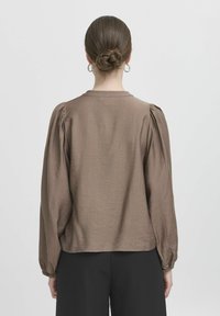 Blusa lunga marrone a maniche lunghe con spalle a sbuffo, tessuto texture, scollatura rotonda e polsini elasticizzati, indossata con pantaloni neri, vista da dietro.