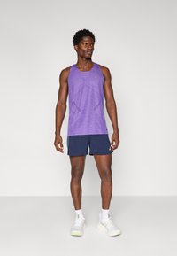 Lila ärmelloses Tanktop mit strukturiertem geometrischem Muster, kombiniert mit navyblauen Shorts und weißen Sneakers mit hellgrünen Akzenten.