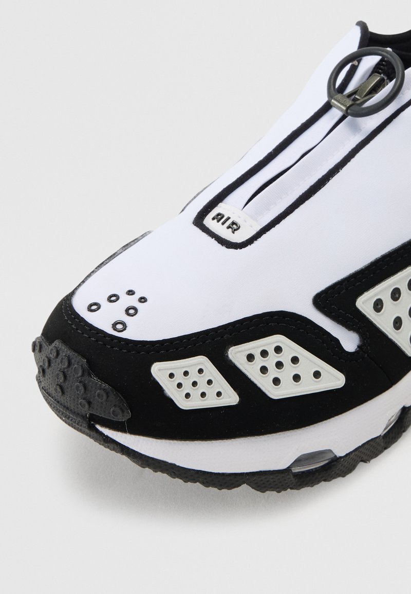 Chaussures de sport avec un schéma de couleurs noir et blanc, dotées d'une fermeture éclair, d'une tige en mesh et d'accents perforés distinctifs.