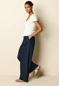 Hvid struktureret top med korte ærmer, parret med marineblå bredbenede bukser og beige flade sandaler, accessoriseret med et ur.