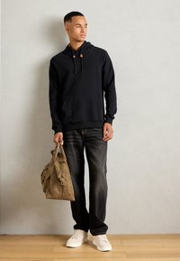 Mann in schwarzer Kapuzenjacke, dunklen Jeans und weißen Sneakers, der eine hellbraune Ledertasche hält und an einer schlichten hellen Wand steht.