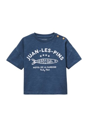 T-shirt bleu bébé à manches courtes, deux boutons en bois sur l'épaule, motif poisson blanc et texte disant Juan-Les-Pins Hotel De La Sardine.