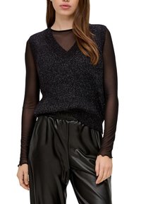 Žena oblečená v černém průhledném topu s dlouhým rukávem, černé bezrukávové pletené vestě a černých kožených kalhotách na bílém pozadí.