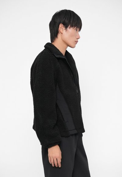 Must have fleece-jakk mustraalse kattega, lukuga ja kõrge kraega. Kerge disain mugava istuvusega, kantud tumesiniste pükste peal.