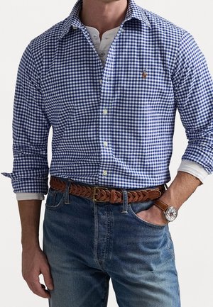 Homme portant une chemise à carreaux bleue et blanche à boutons sur un henley blanc, un jean bleu avec une ceinture en cuir marron tressée, et une montre-bracelet.