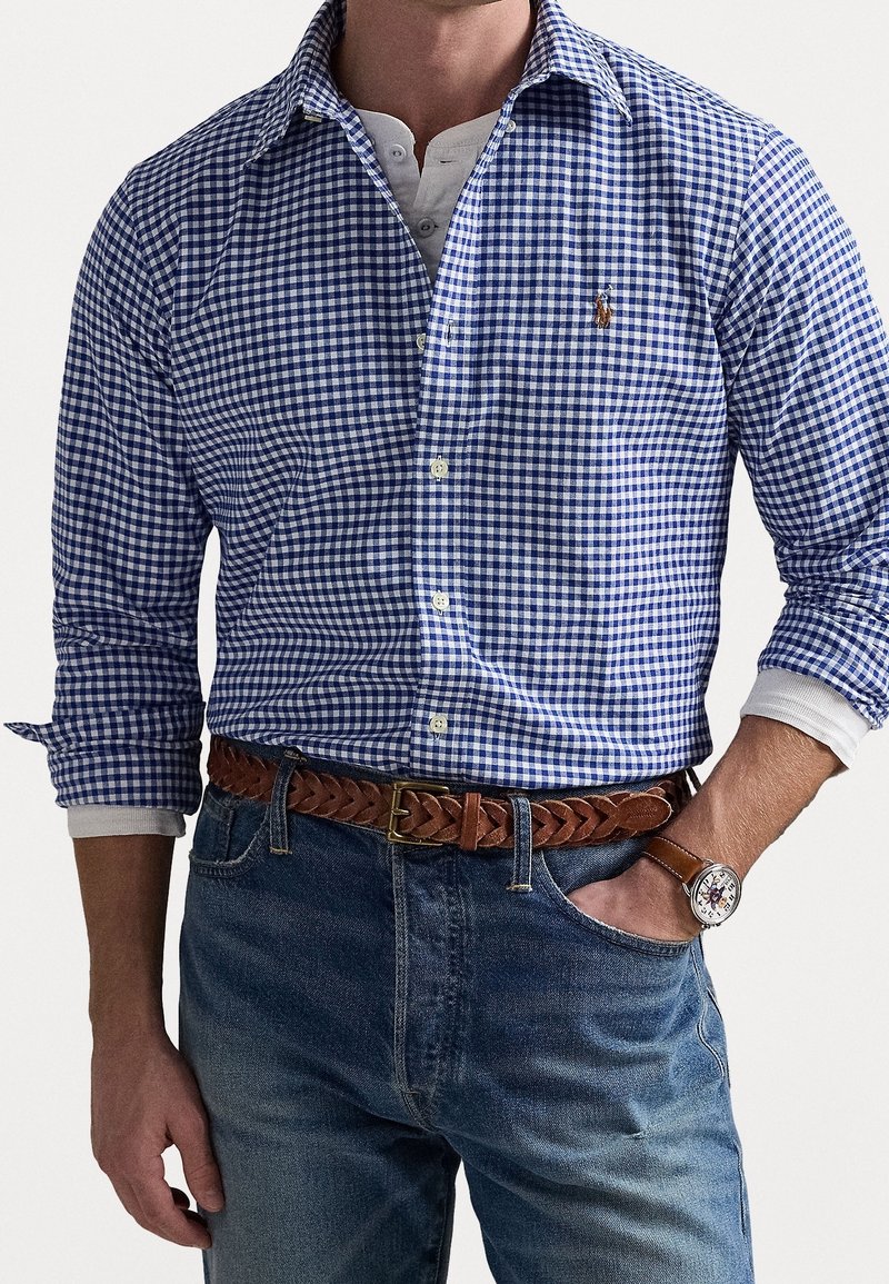 Homme portant une chemise à carreaux bleue et blanche à boutons sur un henley blanc, un jean bleu avec une ceinture en cuir marron tressée, et une montre-bracelet.