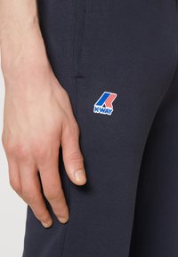 Marineblaue K-Way-Hosen aus einem weichen Stoff, mit schlankem Schnitt und einem kleinen bunten Logo in der Nähe der Tasche. Die Hand ruht auf dem Oberschenkel.