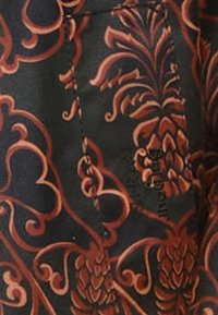 Schwarzer Stoff mit kunstvollen Blumen- und Ananasmustern in satten Brauntönen, sichtbaren Nähten und einem Markenetikett.