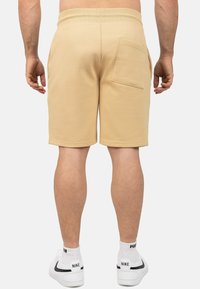 Pantaloni beige con una superficie liscia, cintura elastica e una tasca sul retro. Indossati con scarpe da ginnastica bianche e calzini.