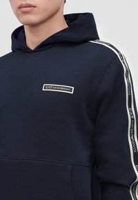 Άνδρας που φοράει μπλε σκούρο φούτερ Emporio Armani με λογότυπο στο στήθος και λωρίδες με την εταιρική σήμανση κατά μήκος των μανικιών.