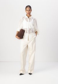 Weiße Spitzenbluse über einem cremefarbenen Shirt, kombiniert mit cremefarbenen Hosen und schwarzen spitz zulaufenden Absätzen. Hält eine braune Leder-Clutch.