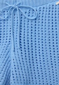 Tejido de punto azul claro con una trama suelta y abierta, con rayas verticales y una cinturilla anudada. Acentuado con pequeños agujeros por toda la superficie.