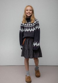 Uśmiechnięta dziewczyna z blond włosami, ubrana w wzorzysty ciemny sweter, kwiecistą spódnicę, szare skarpetki i brązowe buty, stojąca na tle jednolitego tła.