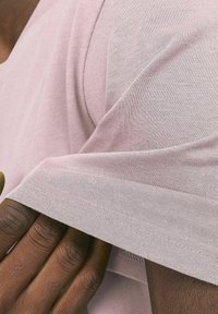 Helles rosa T-Shirt aus Baumwolle, kurze Ärmel und entspannte Passform. Detail einer Hand, die den Ärmel greift, mit sichtbaren Nähten.