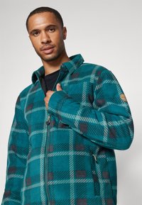 Tepláková bunda v modrozelenom odtieni s kockovaným vzorom. Obsahuje golier, predné zipsové uzatvorenie a vrecká. Mäkko textúra s kontrastnými tmavými akcentmi.