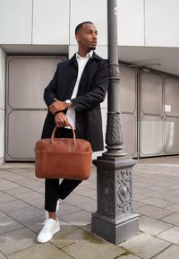 Braune Leder-Laptoptasche mit Obergriffen und Reißverschluss. Glatte Textur und minimalistisches Design, lehnend gegen eine graue Steinsäule.