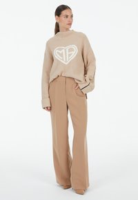 Beige knitted sweater featuring a large white heart logo, paired with light tan wide-leg trousers. Soft texture with long sleeves and ribbed cuffs.