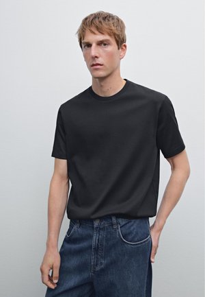 Man met kort blond haar in een eenvoudig zwart t-shirt dat is gestoken in losse donkerblauwe jeans, tegen een lichtgrijze achtergrond.