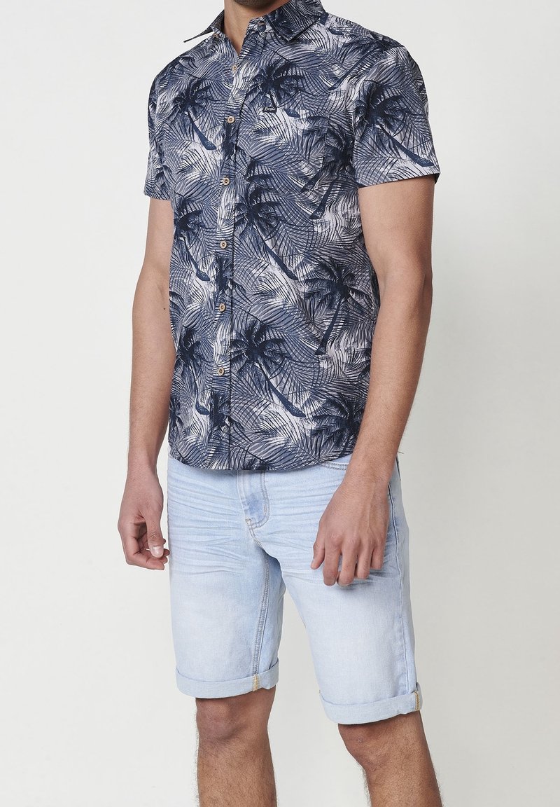 Homme portant une chemise à manches courtes avec un imprimé palmiers bleu foncé et un short en jean bleu clair retroussé, debout devant un fond uni.