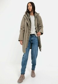 Khaki-Steppjacke mit Kapuze, über einem weiß gestreiften Hemd getragen. kombiniert mit blauen lockeren Jeans und beigen Stiefeln. Glatte Stofftextur.