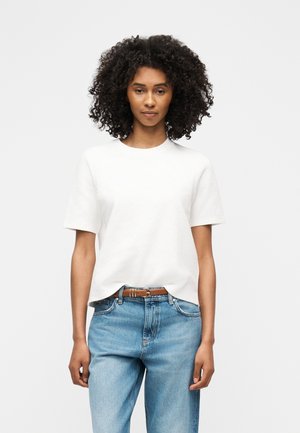 Mlada ženska s kodrastimi lasmi, oblečena v preprosto belo kratko majico, rahlo zataknjeno v svetlo modre kavbojke z visokim pasom in rjavim pasom.