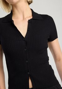 Black ribbed short-sleeve cardigan with a V-neck collar, featuring a button front and side slit detail for a fitted silhouette.