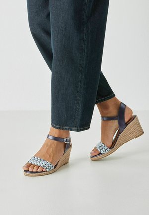 Sandales compensées avec une lanière en tissu tissé bleu et blanc et une lanière de cheville marron foncé. Semelle texturée en liège et design à bout arrondi.