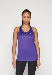 Moteris su pintomis plaukais, dėvinti purpurinį "Nike" be rankovių sportinį marškinėlį ir juodas leggingas, stovinti atsukusi į priekį prieš vienodą foną.