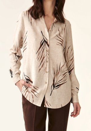 Femme portant un chemisier beige boutonné avec des motifs de feuilles marron et noir, assorti à un pantalon marron foncé, main dans la poche.