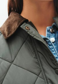 Quilted grøn jakke med en brun kordfløjlskrave, udstyret med lynlås og trykknapper, og en denimskjorte synlig underneden.