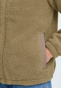 Tamna jakna od flisa s teksturiranom površinom, bočnim džepovima s bež detaljima i zatvaranjem na zip. Mekani materijal i opušteni kroj.