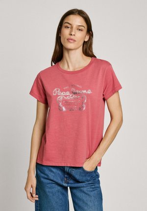 T-shirt en coton rouge clair à manches courtes, col rond, présentant un imprimé graphique argenté avec du texte sur le devant, associé à un jean en denim.