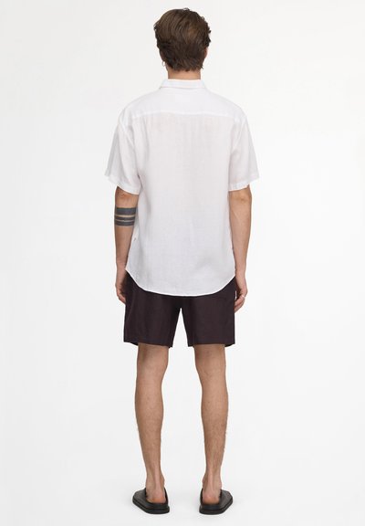 Homme debout, de dos, portant une chemise blanche à manches courtes, un short noir, des sandales noires et un tatouage noir sur le bras gauche.