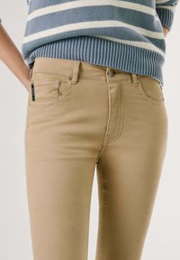 Bež skinny jeans s petimi žepi, sprednjim gumbom in gladkim bombažno mešano tkanino. V paru s sivo prugasto puloverjem.