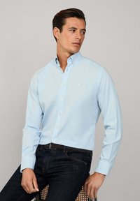 Camicia azzurra chiara con colletto, maniche lunghe e un logo discreto sul petto, abbinata a jeans scuri e una cintura.