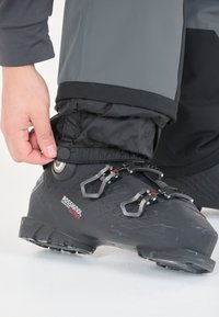 Zwarte skiboot met een gestructureerd oppervlak, metalen clips en rode accenten. Een hand past een zwarte elastische manchet over de laars aan. Een grijze broekspijp is zichtbaar.