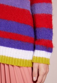 Sweter w paski w kolorach czerwonym, fioletowym i niebieskim z prążkowanym zielono-białym obszyciem. Wykonany z miękkiej, teksturowanej przędzy. Widoczna plisowana różowa spódnica.