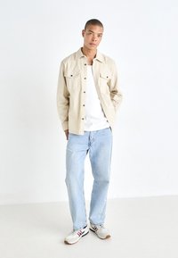 Bež košulja na dugih rukava s dva džepa na prsima, nosi se preko bijele majice, u kombinaciji sa svijetloplinom trapericama i sivim tenisicama s crvenim i plavim detaljima.