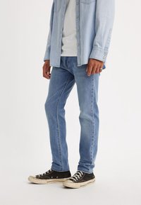 Personne portant un jean en denim bleu clair, des baskets noir et blanc, un t-shirt blanc et une chemise en denim bleu clair avec les manches baissées.