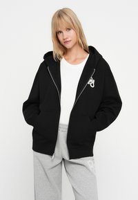 Čierna mikina na zips s prednou vreckom a kapucňou, s bielym logom na hrudi. Nosí sa cez bielu tričko a šedé tepláky.